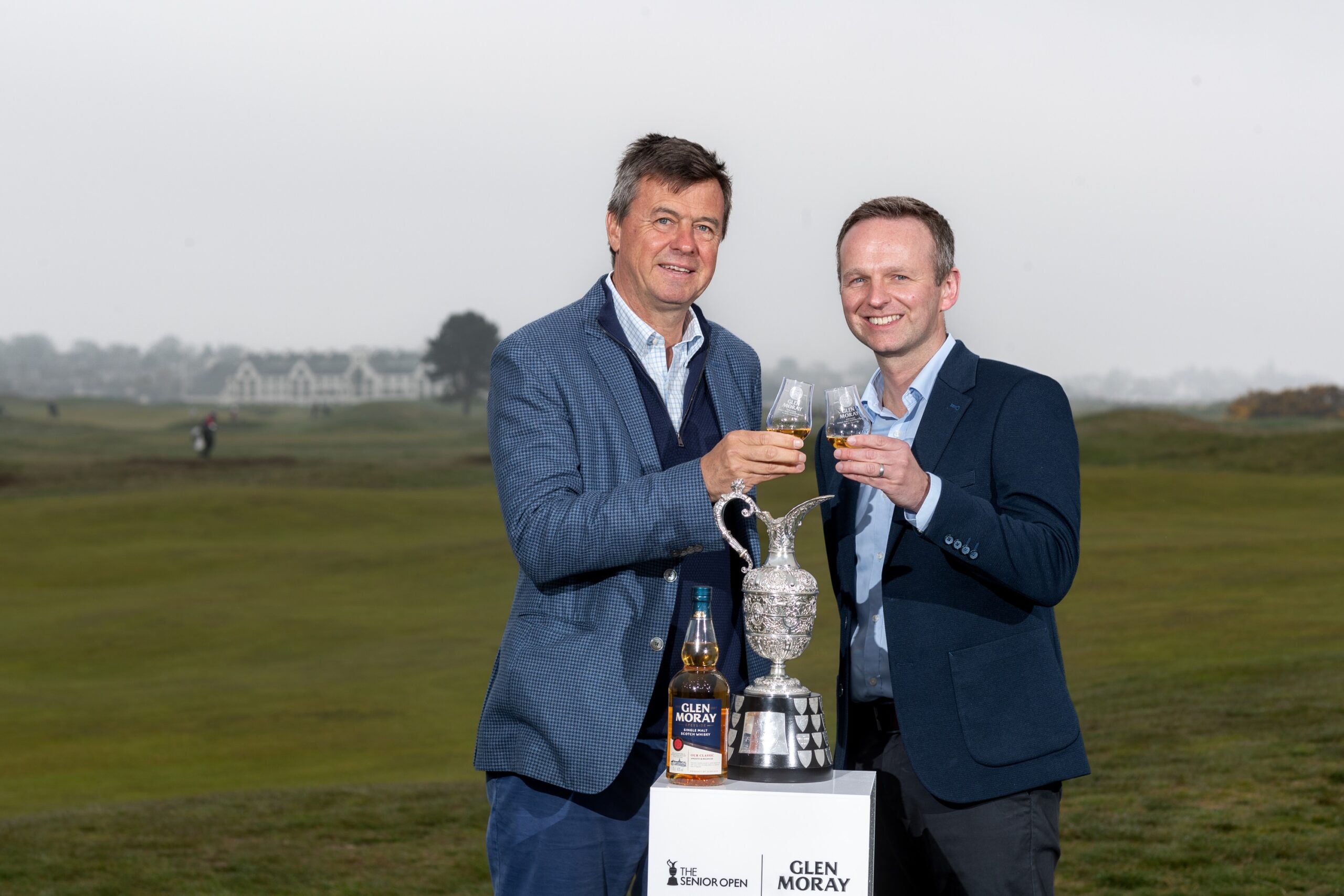 Two officials toasting partnership beside Senior Open trophy