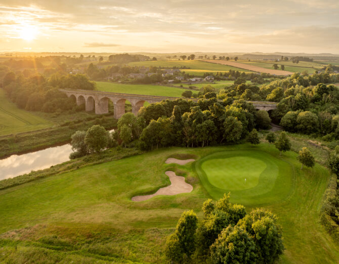 Aerial view of the course with trees and river in the background.